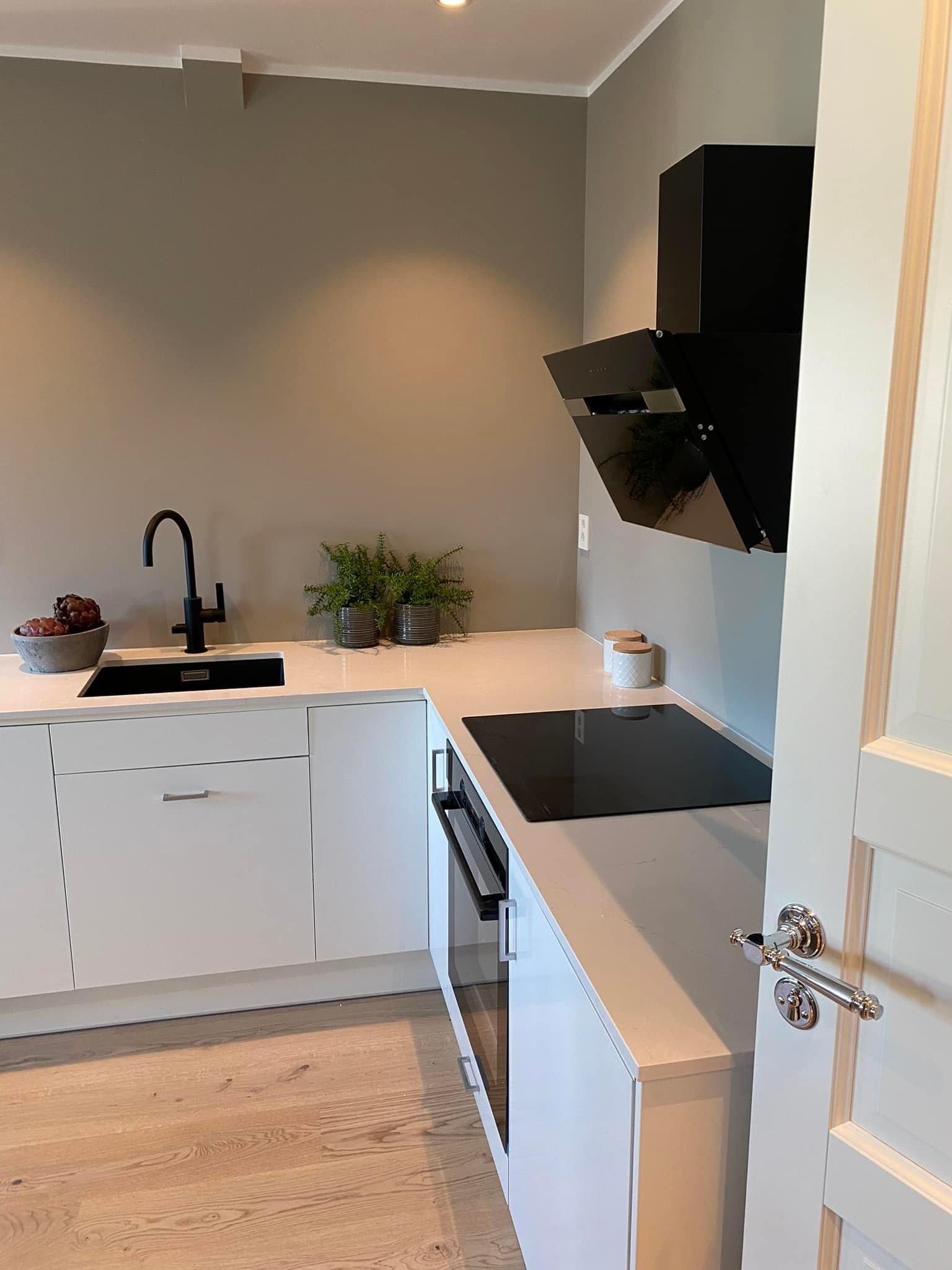Moderne hvitt kjøkken med svarte hvitevarer, gråbeige vegger og lys benkeplate.