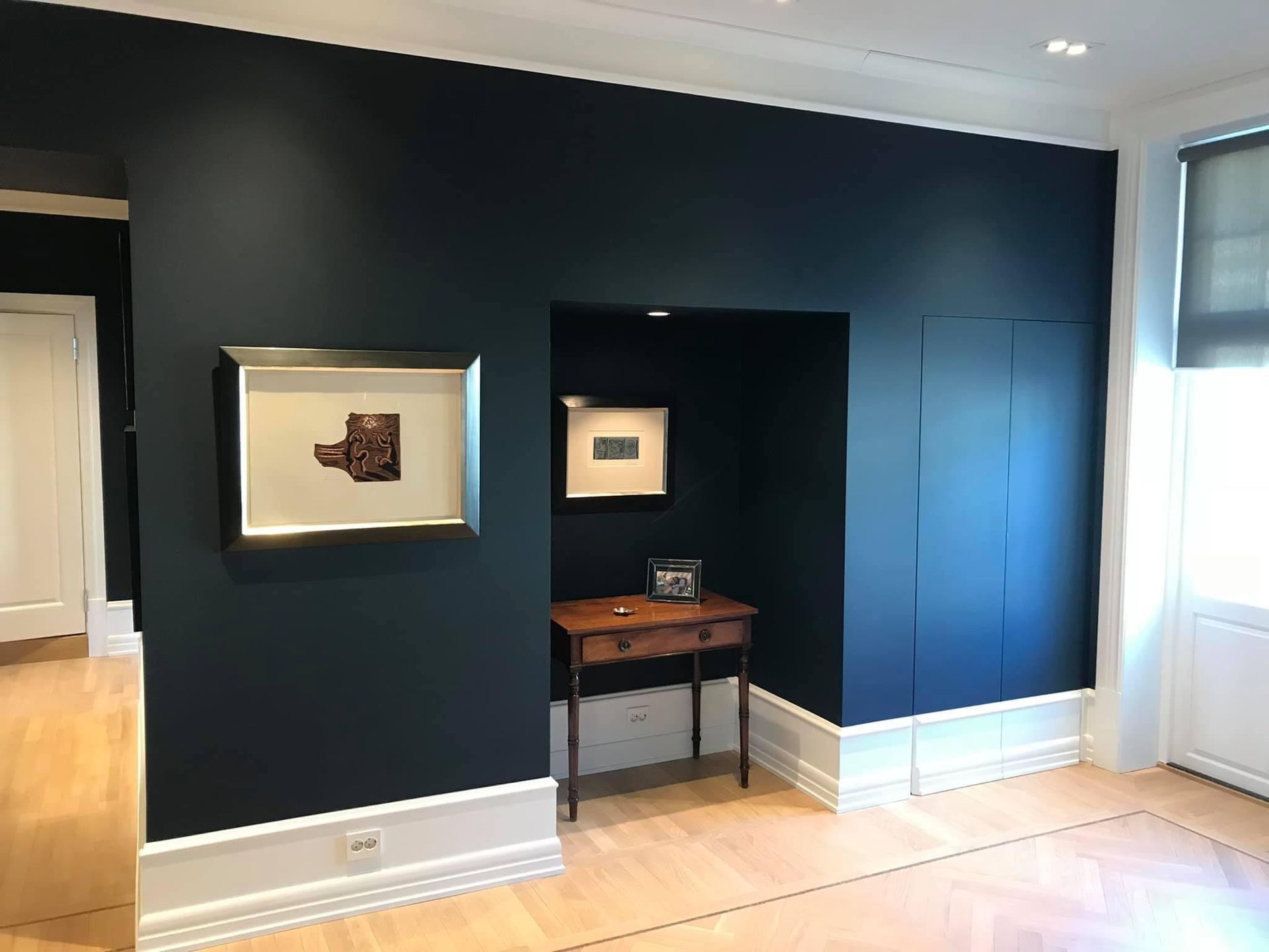 Mørkeblå vegg med kunstverk, lite bord i en innfelt nisje, lyst fiskebensgulv.