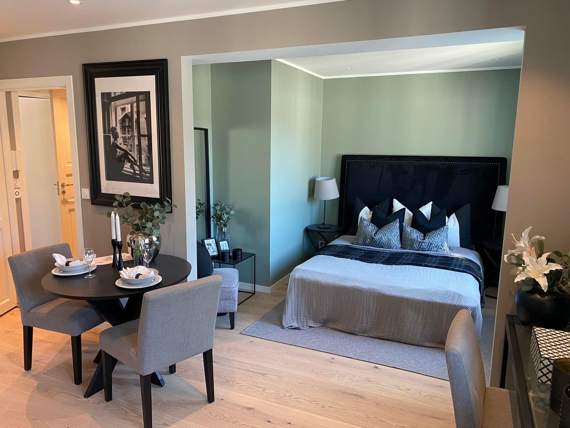 Liten leilighet med soverom og spiseplass. Salviegrønn soveromsvegg, svarte møbler, lite spisebord med stoler.