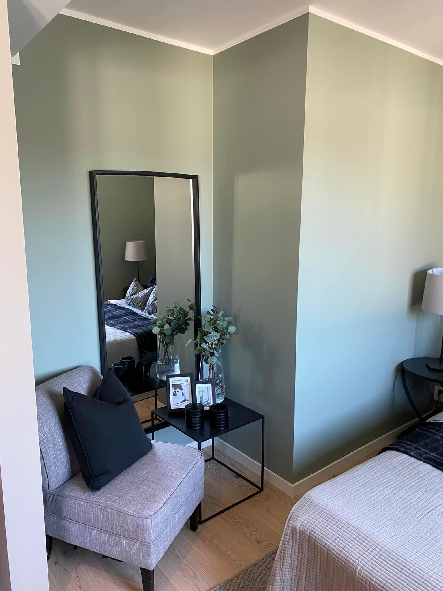 Leilighetshjørne med en salviegrønn vegg, lenestol, svart innrammet speil og et lite sidebord.