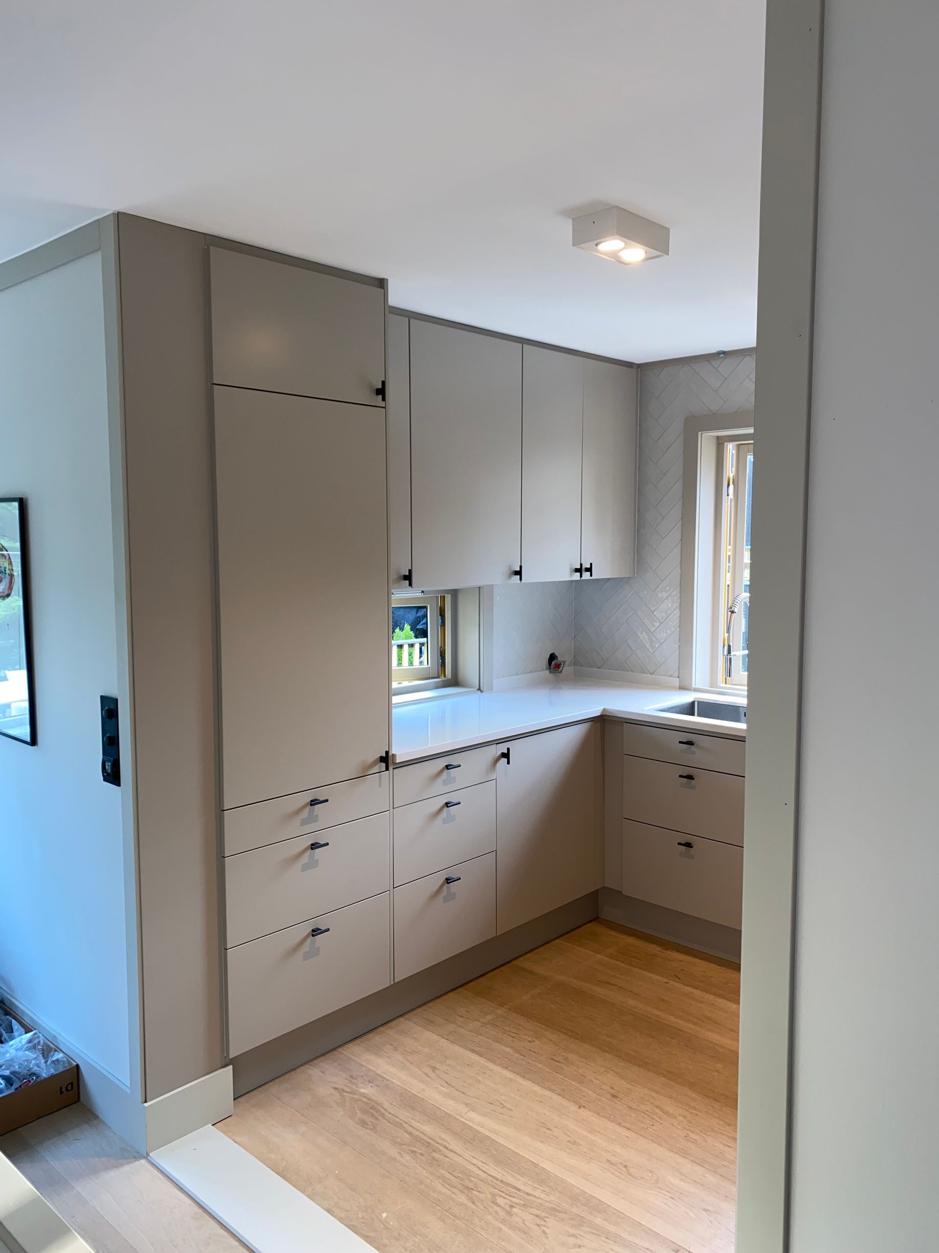 Beige kjøkken med lyst tregulv. Skap med mørke beslag. Vask og vindu synlige.