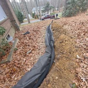 Black silt fence beside a dirt trench covered with fallen leaves in a wooded yard near a house