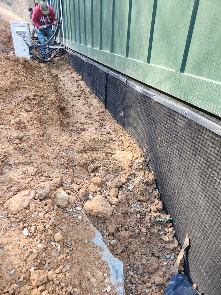 Worker near green building wall by a muddy trench with exposed drainage fabric