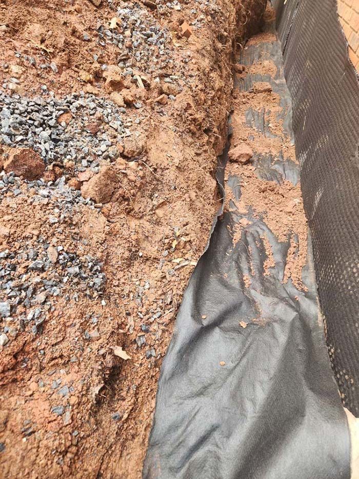 Close-up of a muddy trench beside a black drainage liner and metal mesh wall.