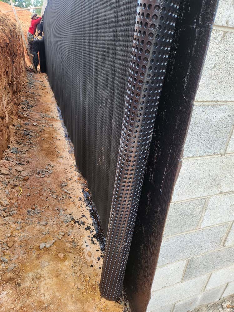 Black drainage mat along cinder block wall beside a dirt trench at a construction site