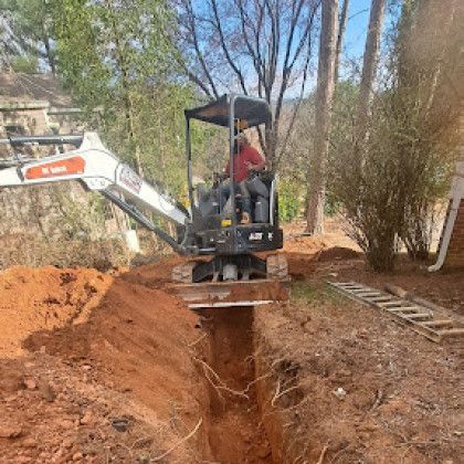 Small excavator digging a deep trench in a wooded dirt yard, with a worker seated in the cab.