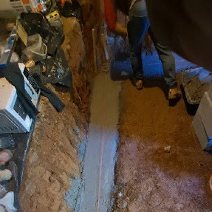 Narrow muddy trench beside a house foundation with exposed pipes and tools.