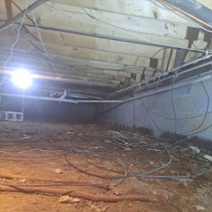 Dim crawlspace or basement with exposed ceiling beams, hanging wires, and dirt floor illuminated by a bright light.