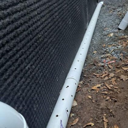 White PVC pipe with drilled holes running along a black wall beside dirt and leaves outdoors