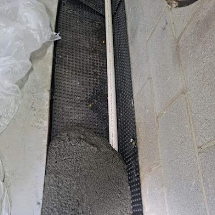 A narrow trench with black mesh liner and gray concrete wall blocks beside a white pipe.