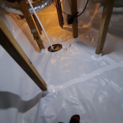Plastic sheeting covering floor joists in a crawlspace with a white pipe and warm work light