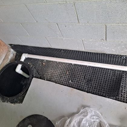 White PVC pipe runs along cinderblock wall beside a black plastic tub and rubber mat on concrete floor