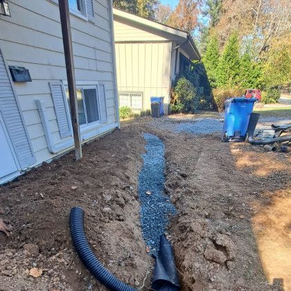 Backyard trench with gravel drainage line beside a house, construction materials and blue trash bins nearby