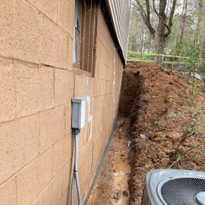 House wall with electrical box beside a muddy trench and dirt pile in a wooded yard