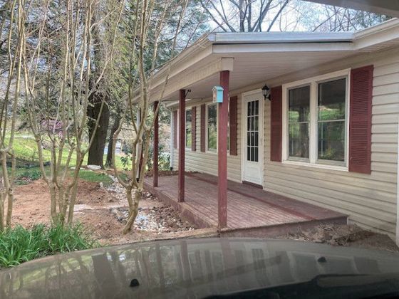 Front porch of a beige house with red shutters and railing, viewed from a driveway, surrounded by trees.