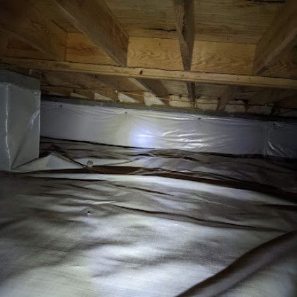 Inside a crawlspace, plastic vapor barrier covers the floor beneath wooden joists, lit by a flashlight.