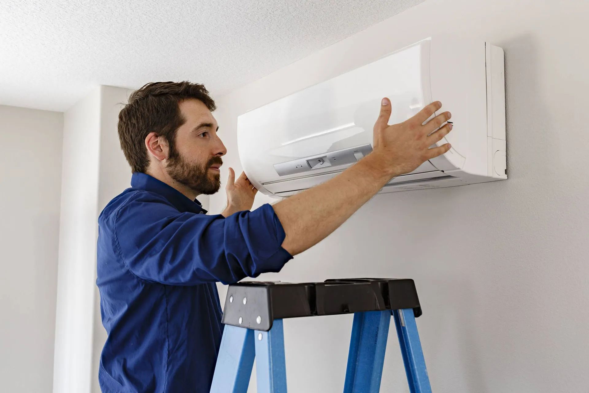 Technician installing a white wall-mounted air conditioner on a blue ladder