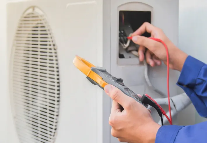 Hands using a multimeter to test wires inside a wall outlet box beside a vent.