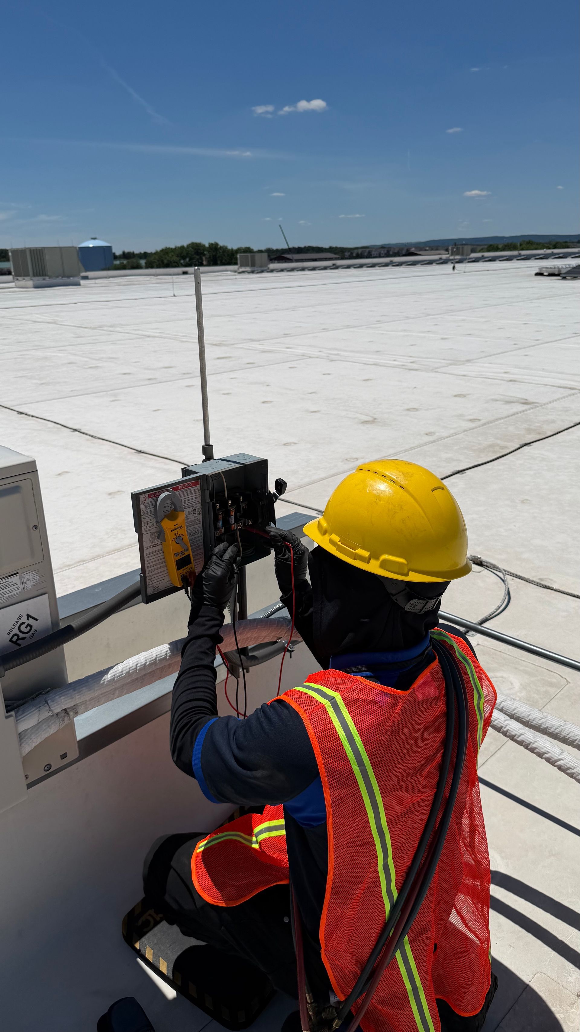 Worker in yellow hard hat and orange vest operating equipment on a white rooftop under a clear sky