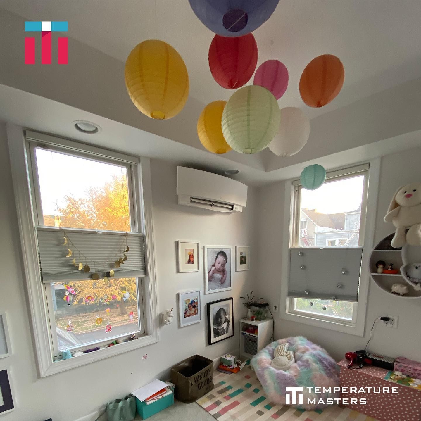 Bright bedroom with colorful hanging lanterns, framed wall art, windows, and a small desk area.