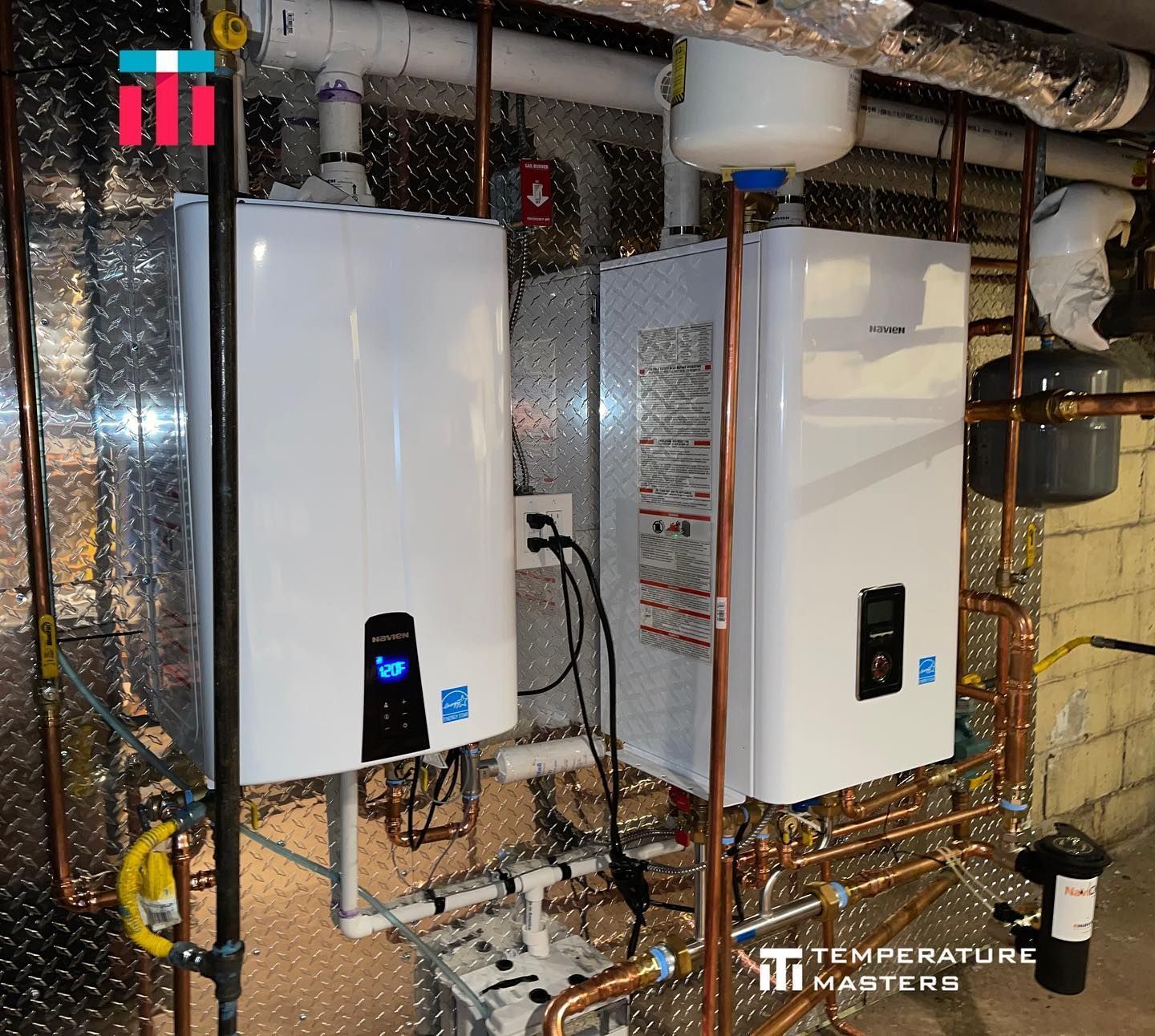 Two white wall-mounted tankless water heaters in a utility room with exposed pipes and foil insulation.