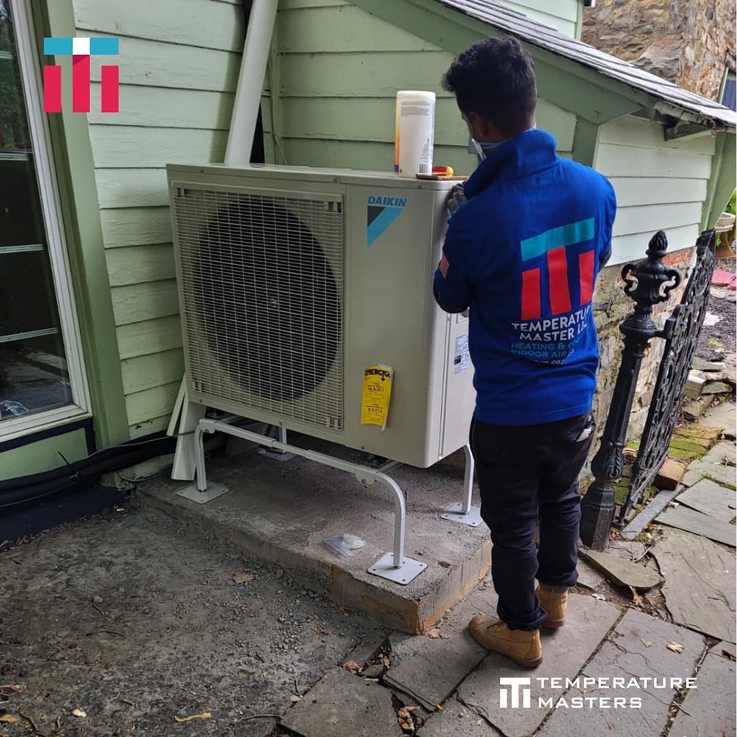 Technician inspecting an outdoor HVAC unit beside a green house wall.