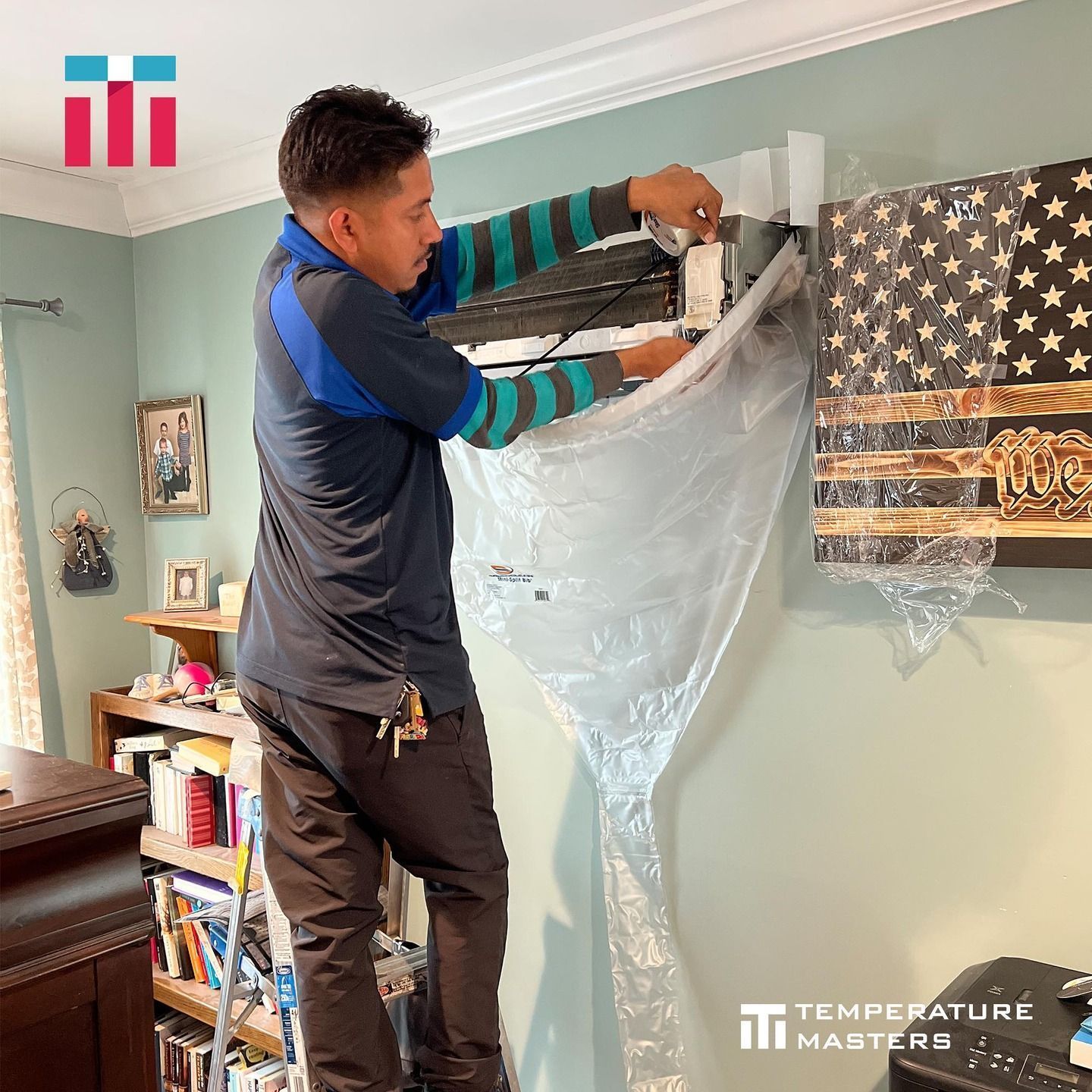 Man hanging clear plastic sheeting over a wall-mounted TV in a living room