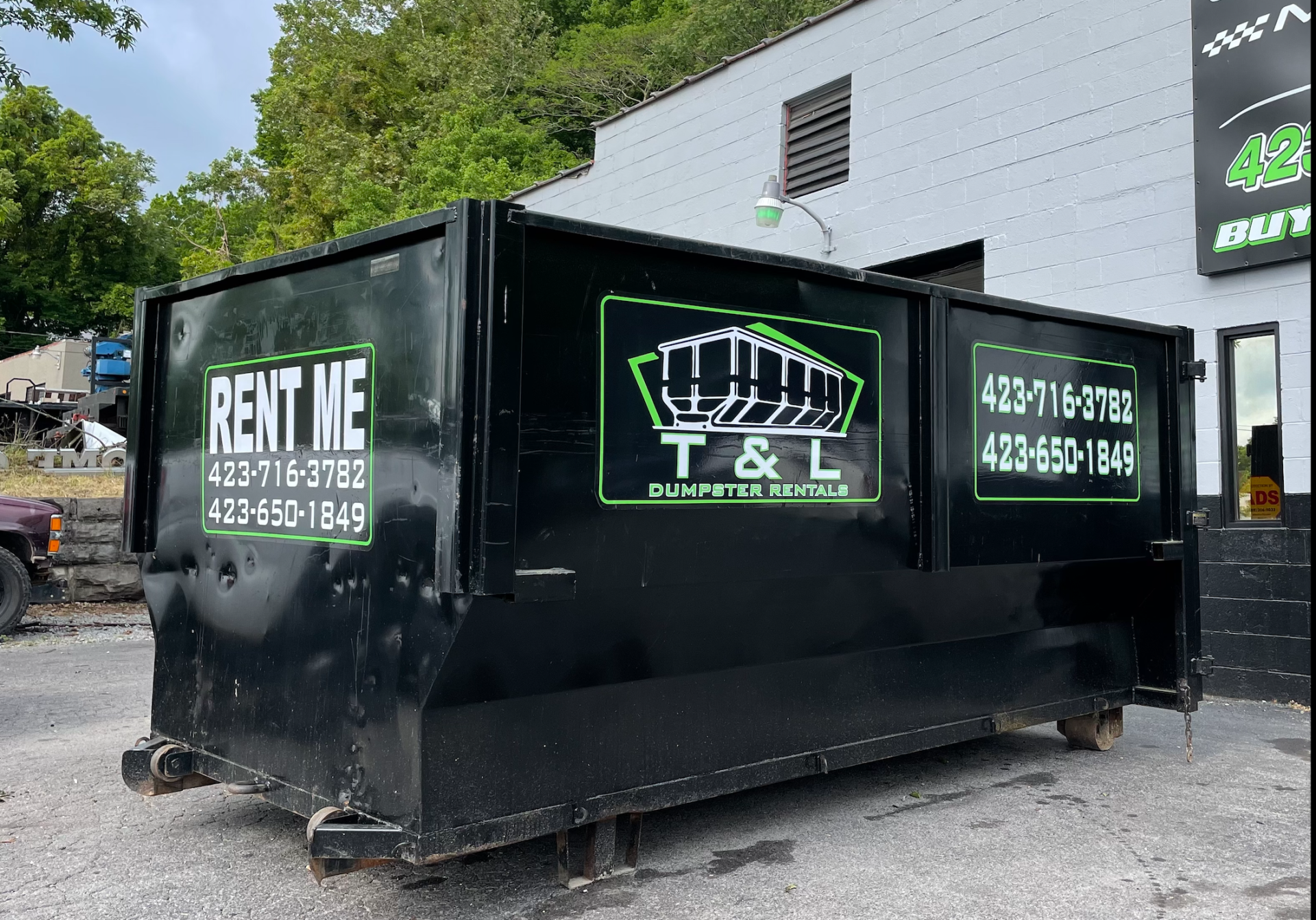 A black dumpster is parked in front of a building that says rent me t & l.