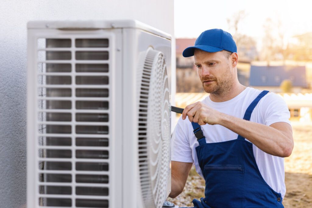 Technician in blue overalls and cap inspecting an outdoor air conditioner unit with a screwdriver