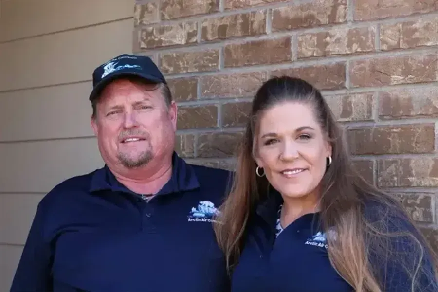 Two people in navy shirts and caps standing in front of a brick wall, smiling.
