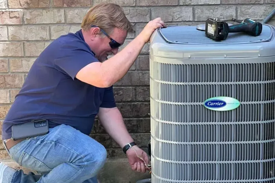 Technician crouches beside outdoor air conditioner unit, inspecting it against a brick wall.