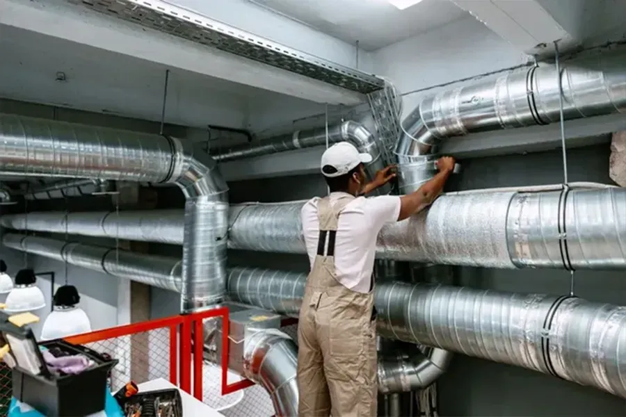 Workers installing large silver ventilation ducts in an industrial interior, one on a ladder inspecting pipes.