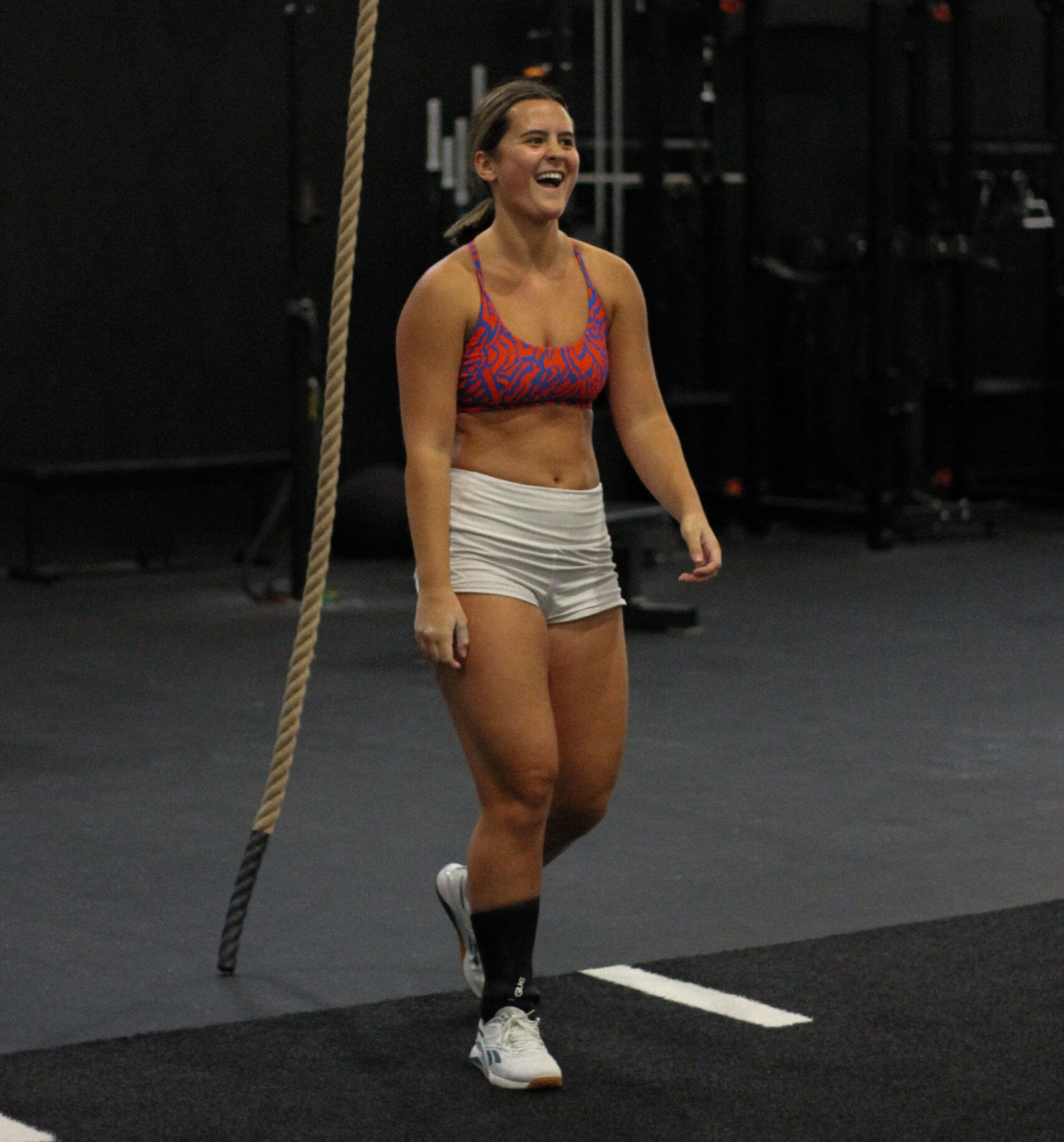 Woman in sports bra and shorts smiles in a gym, rope visible.