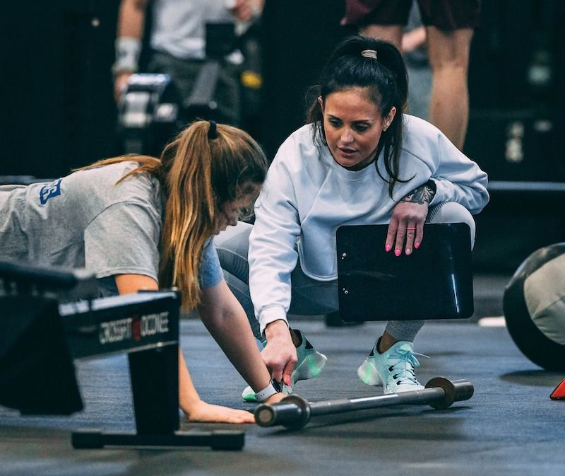 Woman coach helping another woman with a barbell at a gym. Both are focused and attentive.