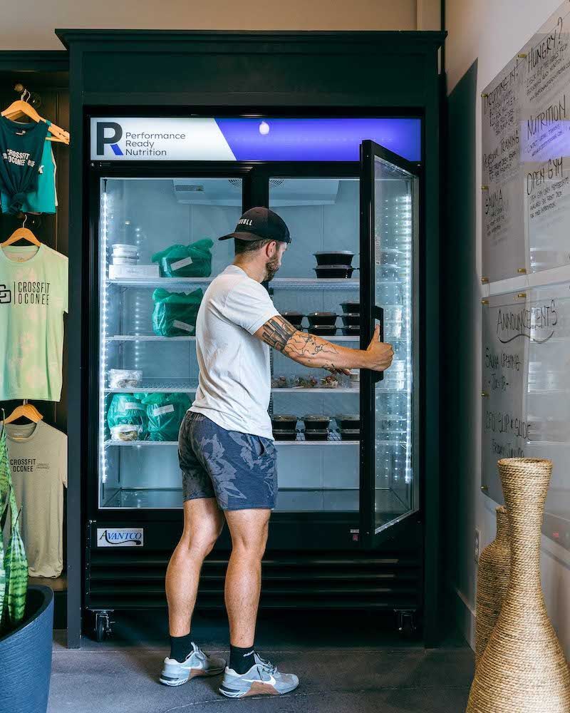 Man opening a refrigerated food display case. Inside are packaged meals. Shirts hang on the left.