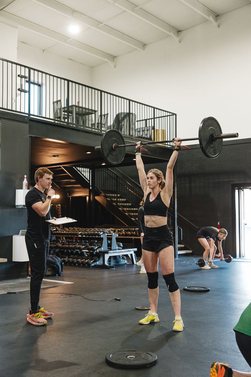 Woman overhead presses a barbell in a gym, watched by a coach. Other people workout in the background.