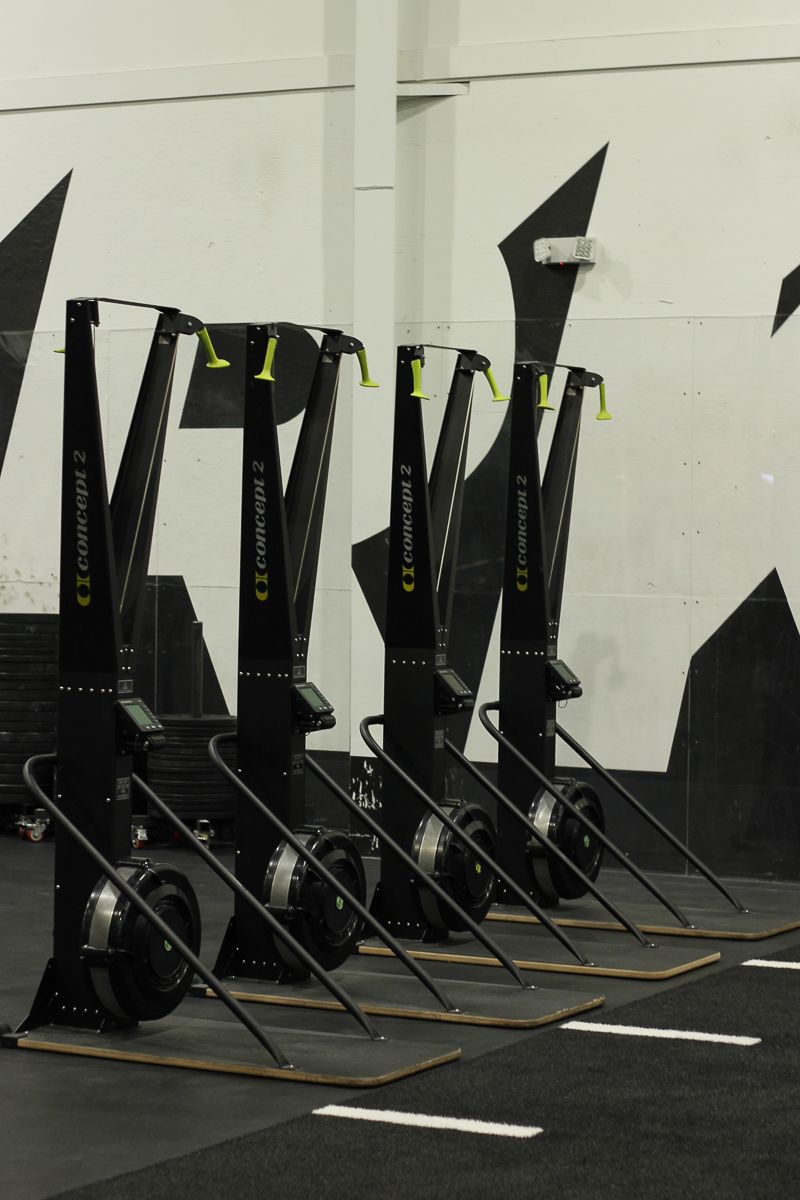 Rowing machines lined up in a gym, black and yellow accents, on wooden platforms.