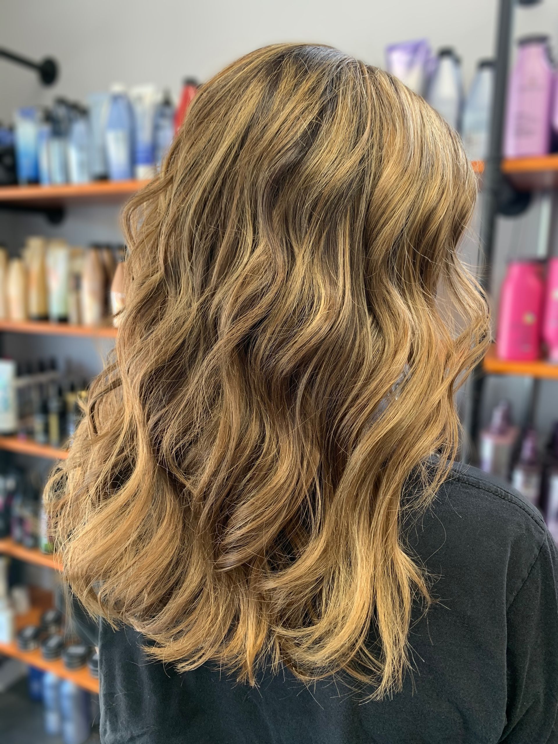 Back view of shoulder-length, wavy, highlighted brown hair in a salon setting with product shelves in the background.