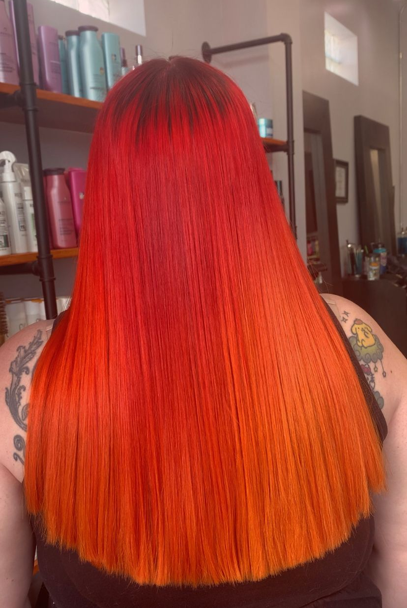 Long hair with a vibrant ombre transition from deep red at the roots to bright orange at the ends, inside a salon.