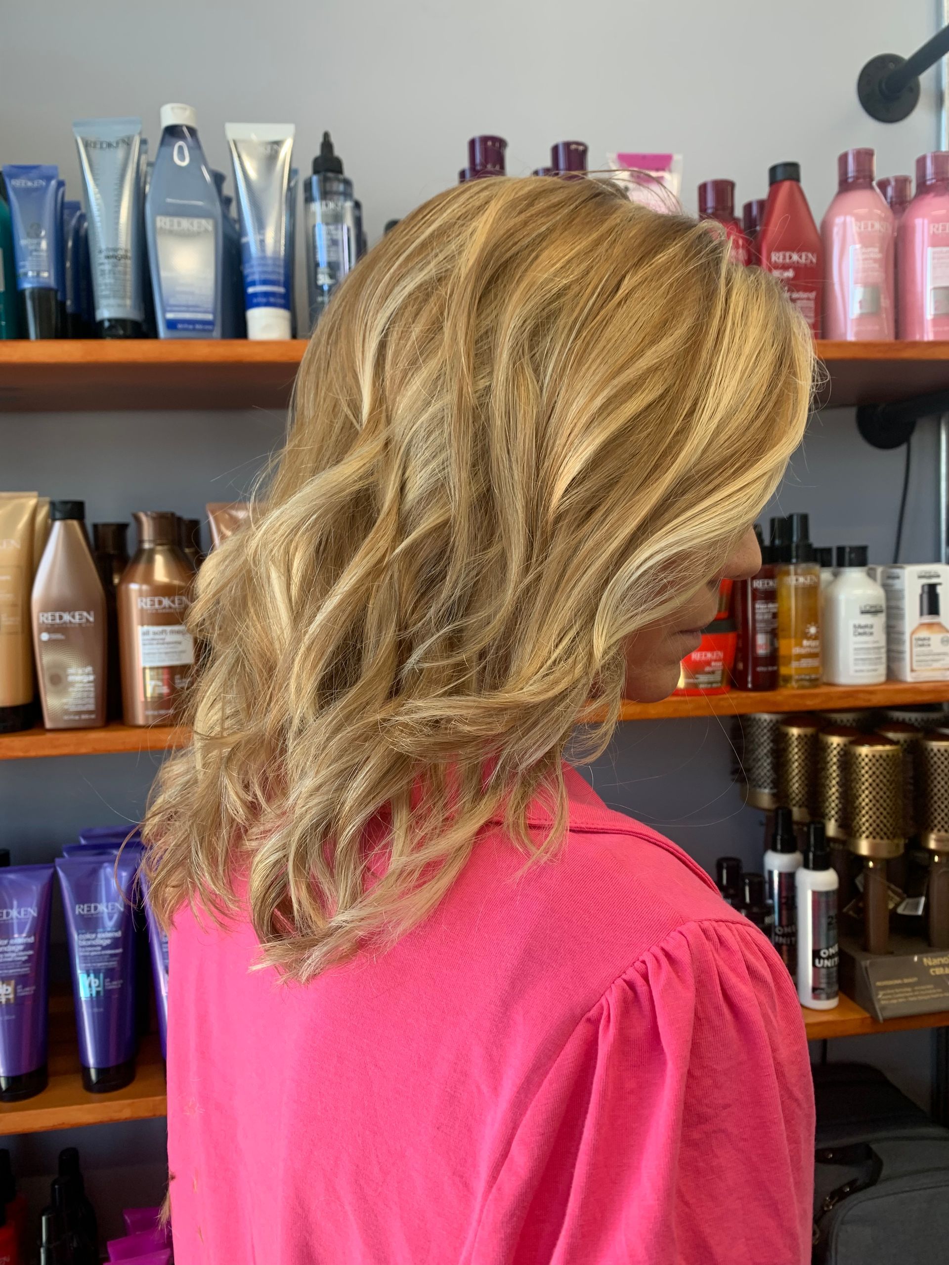 A person with wavy, golden-blonde hair stands in a salon in front of shelves stocked with various hair care products.