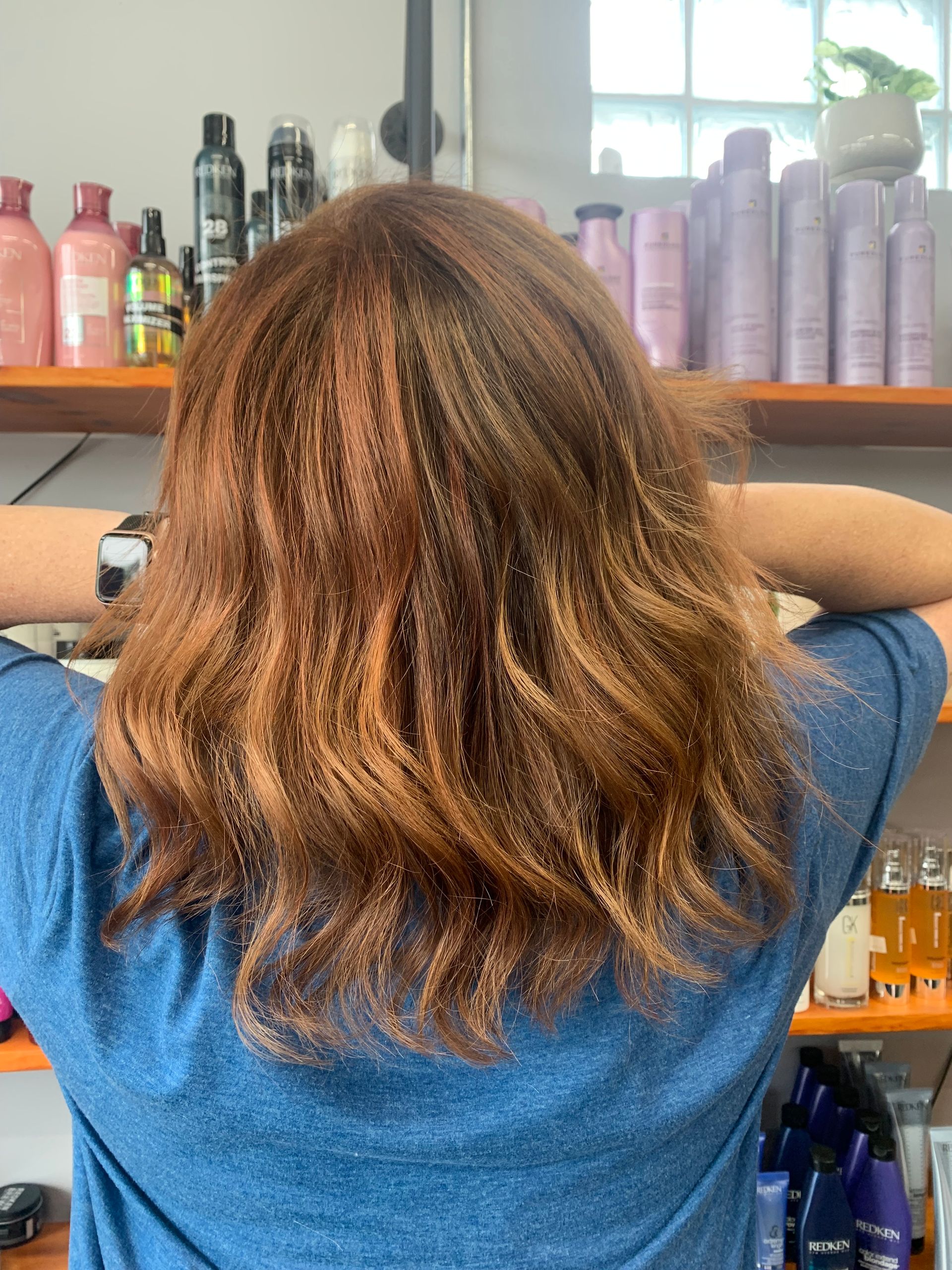 A person with shoulder-length, wavy, reddish-brown hair stands in front of hair salon shelves filled with product bottles.