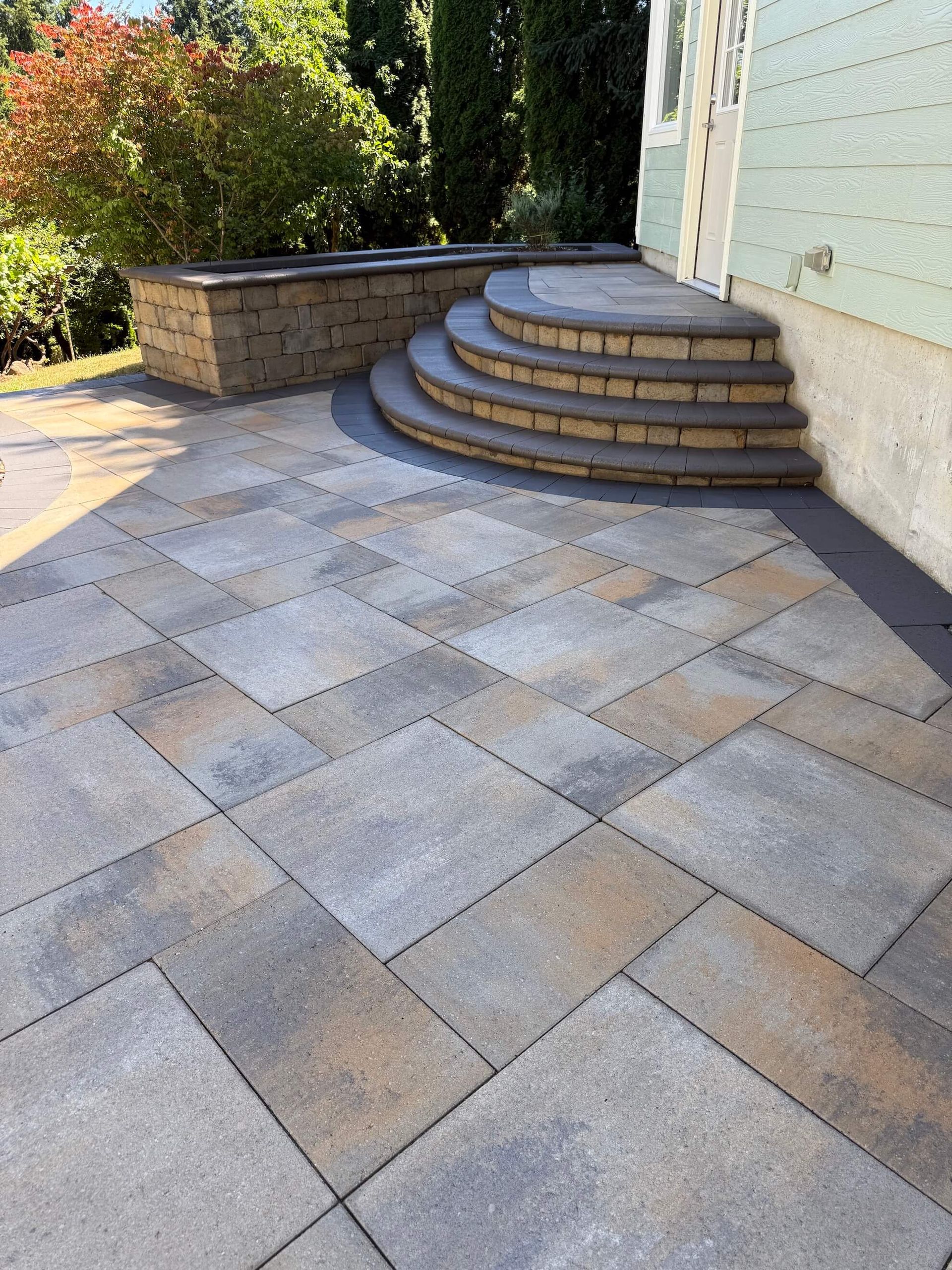 A curved three-step stone staircase leads to a light-colored house exterior, beside a paved stone patio and a brick wall.