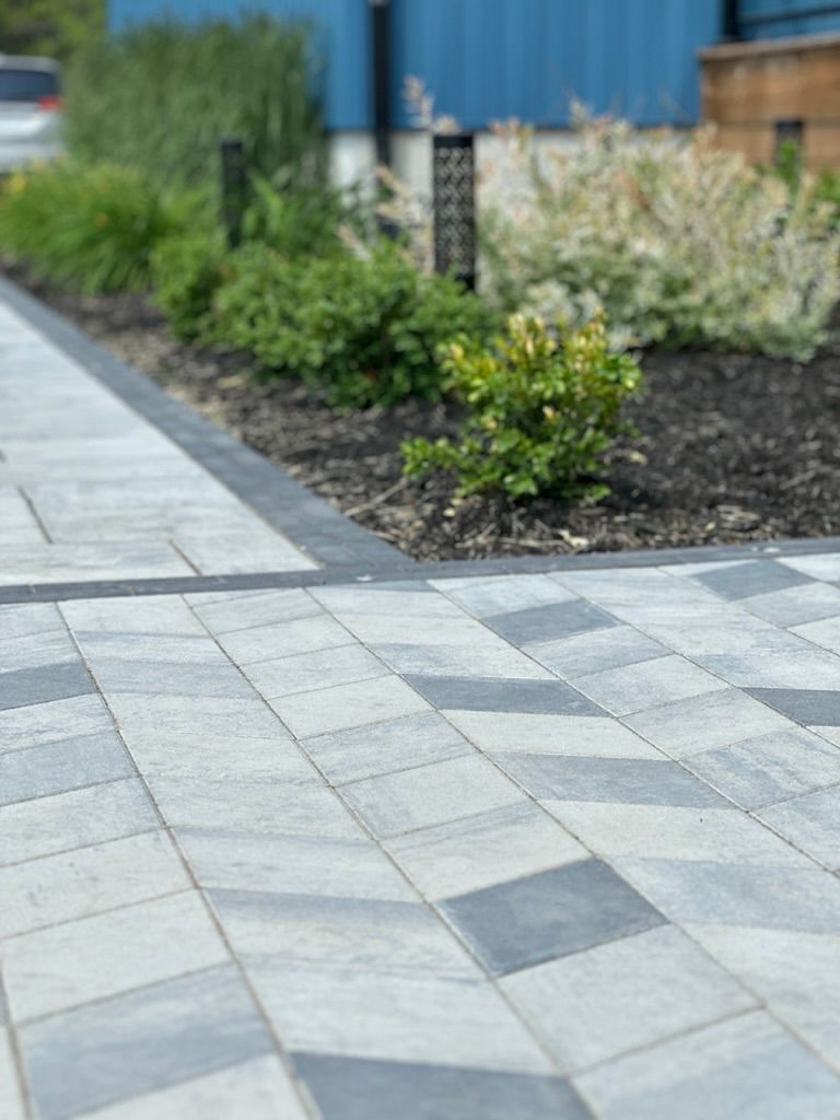 Brick pavers leading to a garden bed with lush greenery, against a blue building backdrop.