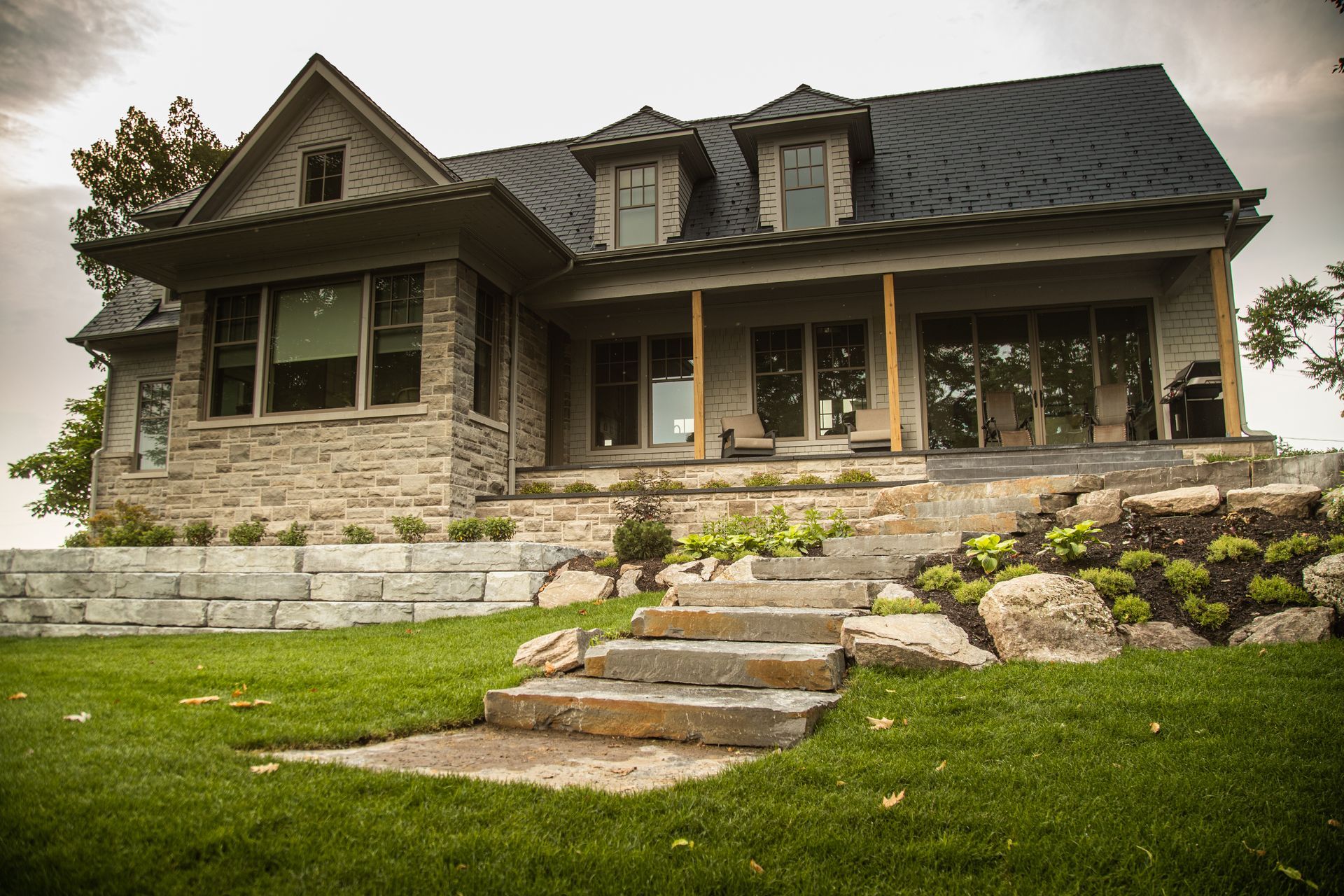 Stone house with black roof, large windows, and a porch, set on a grassy hill with stone steps.