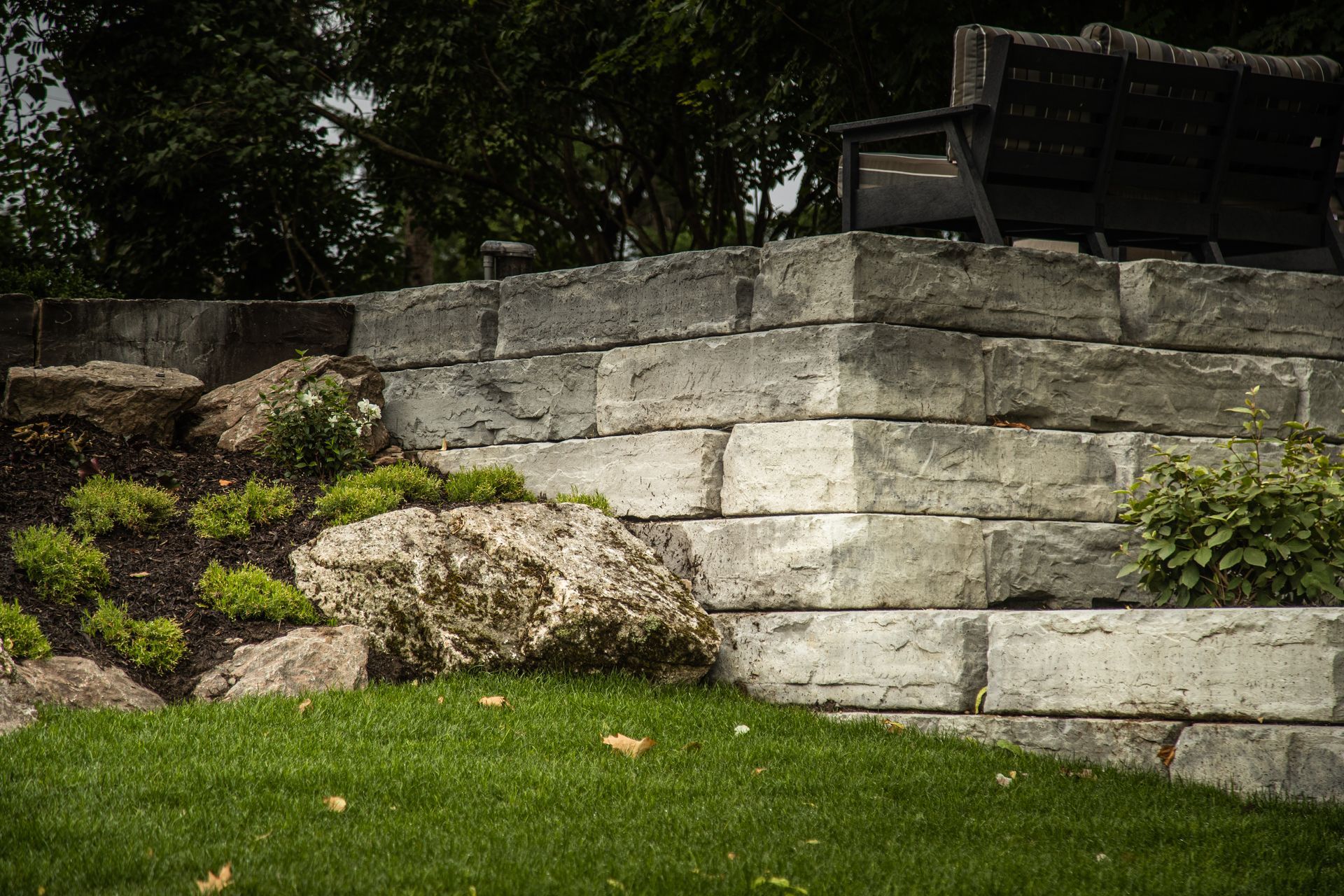 Stone retaining wall with a grassy lawn in front and greenery.