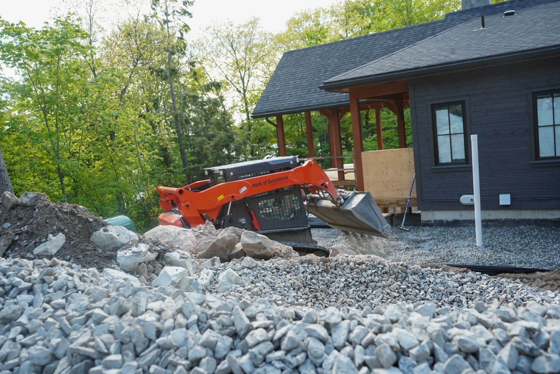 Orange skid steer loader moving gravel near a house with a dark facade, trees in the background.