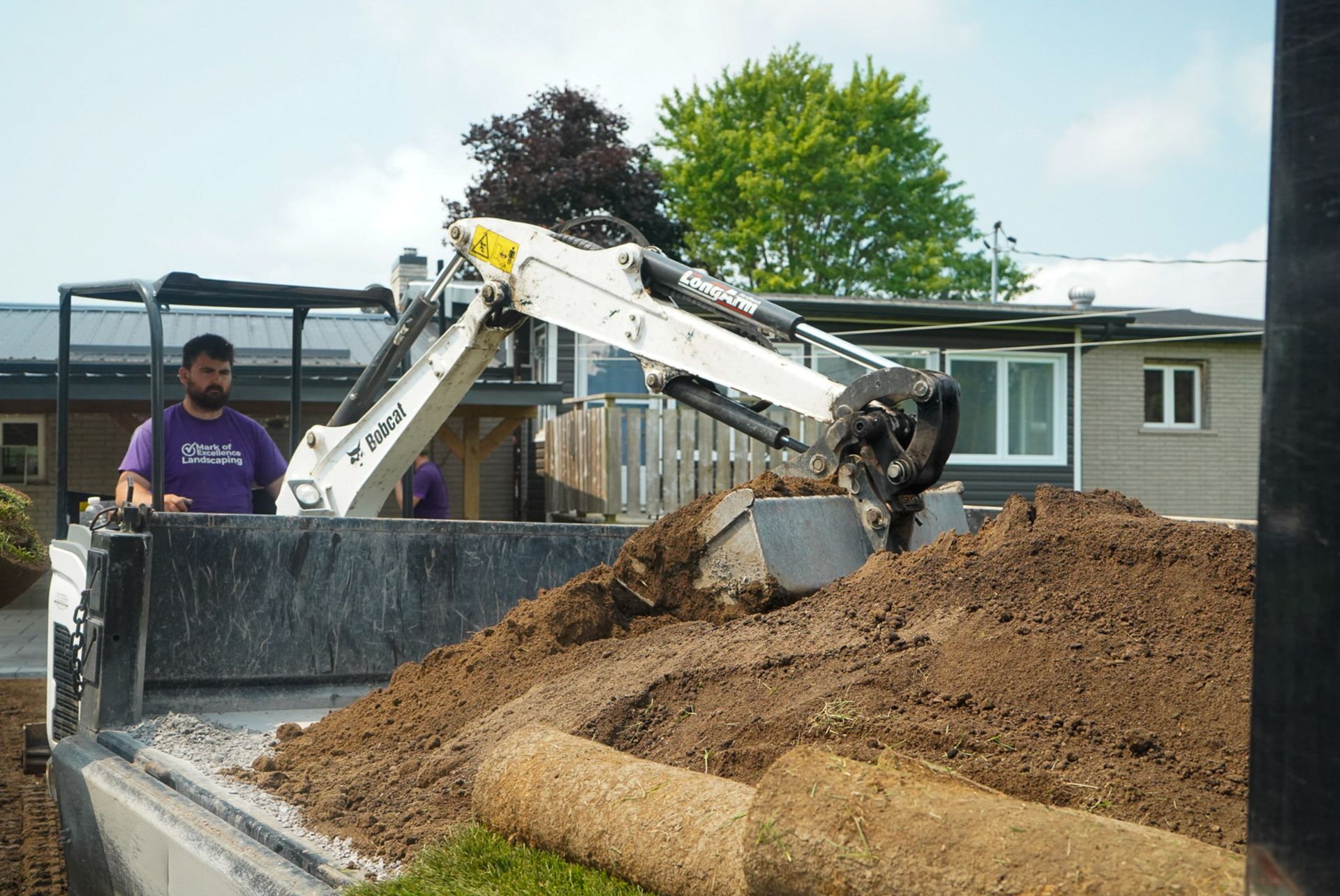 A Bobcat excavator loads soil into a truck, a man watches. Outdoors. Sunny.
