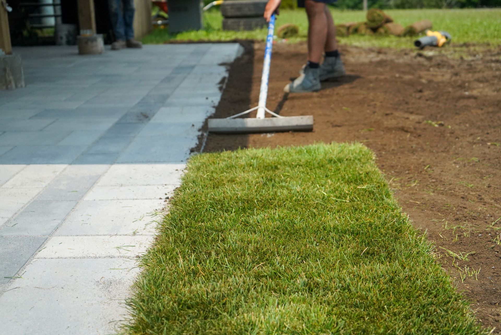 Person raking dirt next to new sod bordering a paved patio.