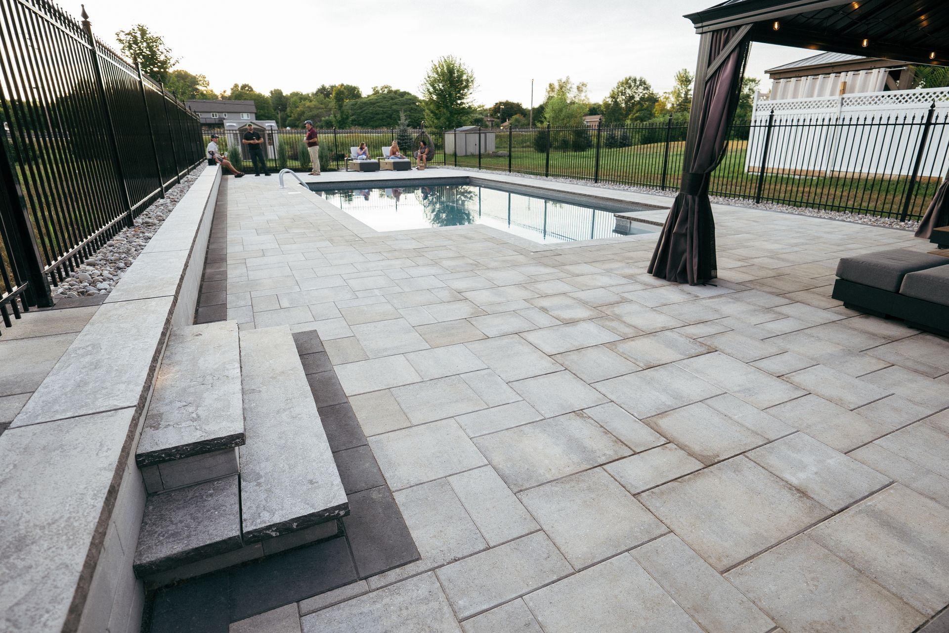 Swimming pool and patio with gray pavers, steps, and black fence.