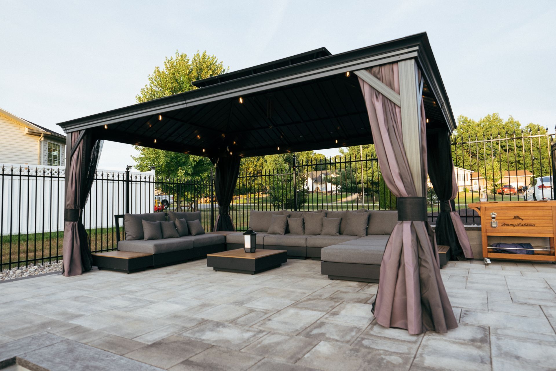 Gazebo with gray couches on a paved patio, black curtains, and string lights.